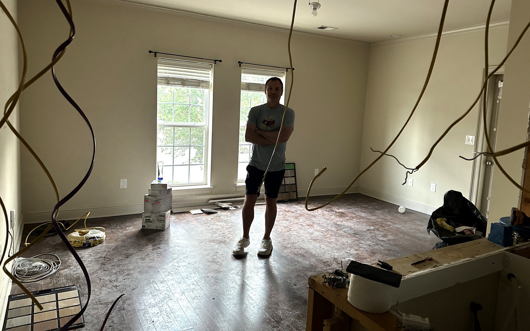 The same room mid-renovation: bare subfloor, wiring hanging from the ceiling, boxes of fixtures, and Lance standing in the middle of it.
