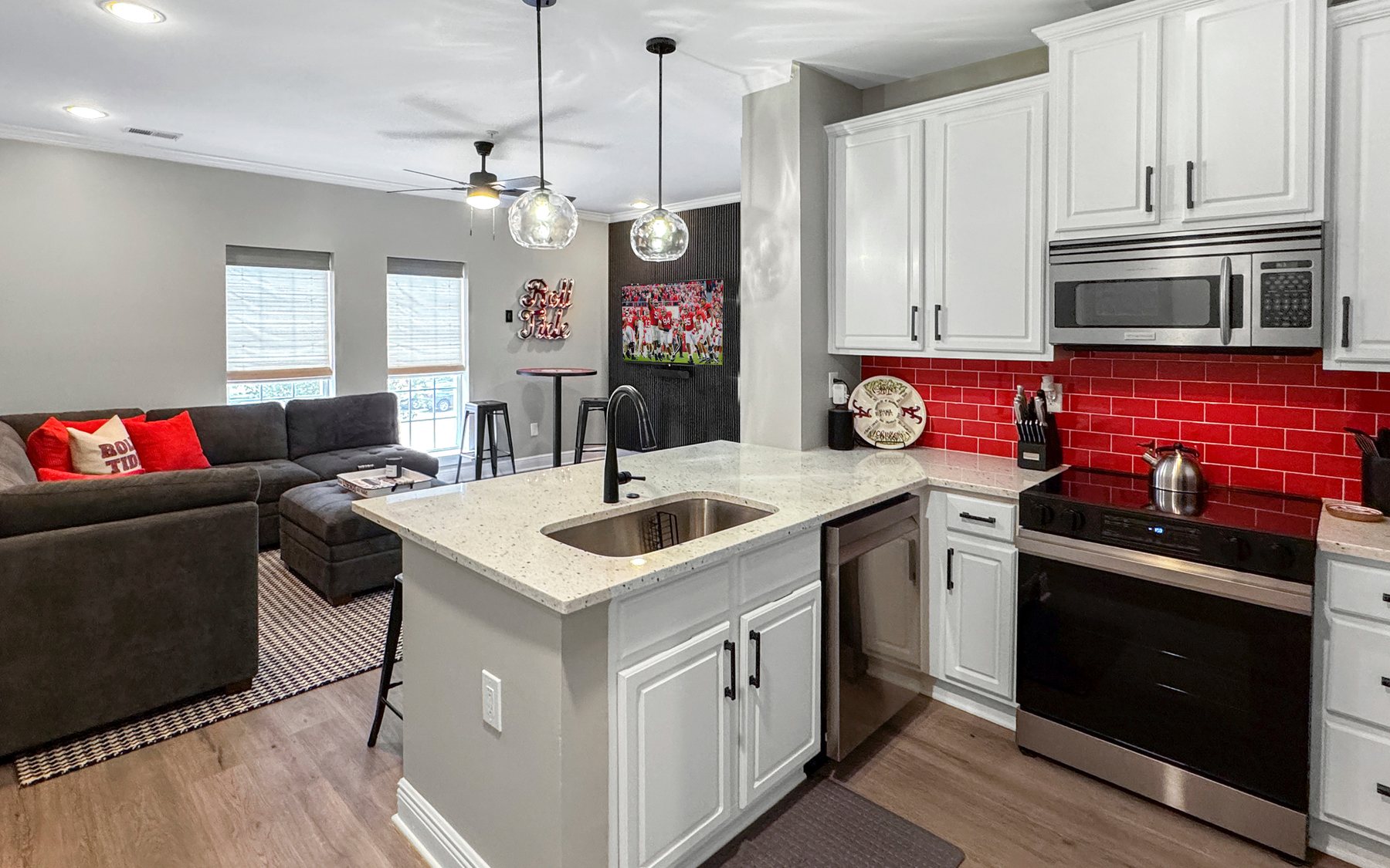The finished room: open kitchen with a red tile backsplash, island, sectional, houndstooth rug and a game on the television.